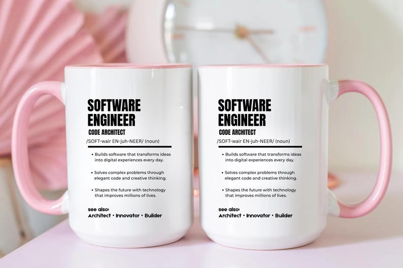 K&ouml;nnte beinhalten: Zwei wei&szlig;e Keramiktassen mit hellrosa Henkeln und Innenseiten. Jede Tasse zeigt schwarzen Text, der einen Softwareentwickler als Code-Architekten definiert, inklusive Aufz&auml;hlungspunkten &uuml;ber Softwareentwicklung und technologische Innovation.