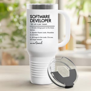 Może przedstawiać: Biały kubek podróżny z uchwytem i przezroczystą pokrywką. Kubek zawiera tekst "SOFTWARE DEVELOPER" z definicją i humorystycznymi stwierdzeniami na temat tworzenia oprogramowania. Zawiera również imię "Daniel".