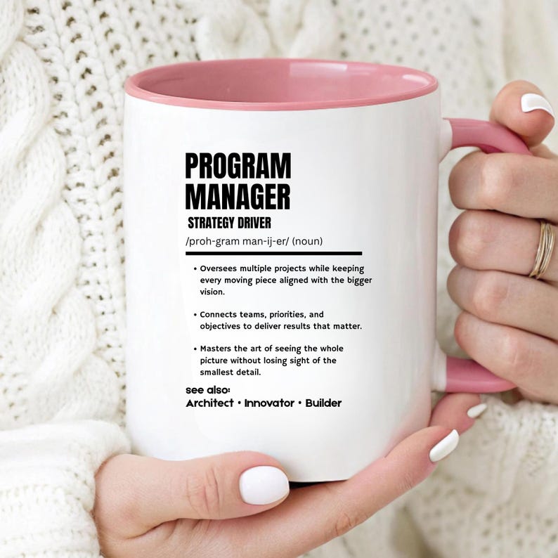 May include: A white ceramic mug with a pink interior and handle, featuring black printed text that defines a Program Manager as a strategy driver, including bulleted job responsibilities and the terms Architect, Innovator, and Builder.