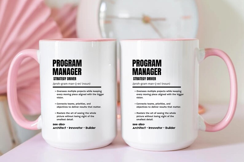 May include: Two white ceramic mugs with light pink handles and interiors. Each mug features black text defining a Program Manager as a strategy driver, including bullet points about overseeing projects, connecting teams, and mastering details.