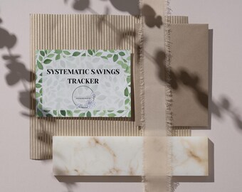 May include: A flat lay featuring a "Systematic Savings Tracker" card with a floral design, surrounded by textured paper, a marble block, and a ribbon. The card has a green leaf border.