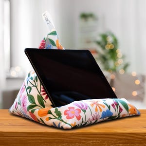 Peut inclure: Un coussin pour tablette à imprimé floral en forme triangulaire, soutenant une tablette noire. Le coussin présente un fond blanc avec des fleurs colorées et des feuilles vertes. Une petite étiquette blanche avec le texte "LARICA" est visible.