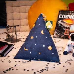May include: A blue triangular cushion with a celestial pattern of stars, moons, and suns. The cushion is on a white surface with silver sequins.