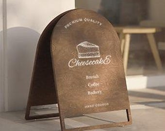 May include: A brown wooden A-frame sign with a white chalk drawing of a slice of cheesecake and the text "Cheesecake" in cursive. The sign also has the text "Premium Quality" at the top and "Brunch, Coffee, Bakery, Hand Cooked" at the bottom.