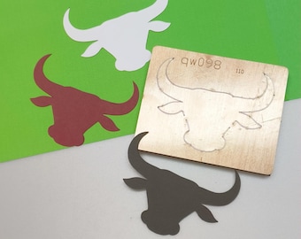 May include: A collection of bull head cutouts in white, burgundy, and dark gray, along with a wooden die-cut template. The template has the text "qw098" and "110" etched into it. The cutouts are on a green and white background.
