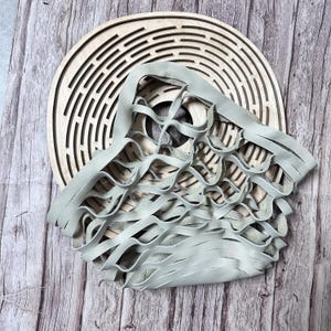 Może przedstawiać: Jasnoszara skórzana torebka z falistym, frędzelkowym wzorem, nałożona na okrągłe drewniane dzieło sztuki z koncentrycznymi rowkami. Tekstura torby kontrastuje z gładkim drewnem.