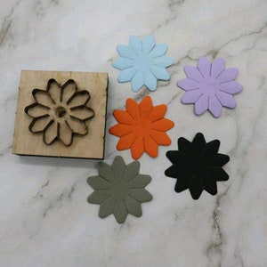 May include: A wooden flower-shaped die-cut tool with a metal cutting edge, next to six colorful flower cutouts in blue, orange, purple, black, and gray. The die-cut tool is square and the cutouts are in a variety of colors.