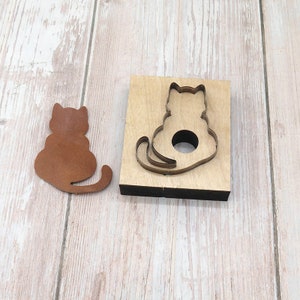Könnte beinhalten: Ein Holz-Stempel mit einer ausgeschnittenen Katzenform. Neben dem Stempel liegt ein braunes Leder-Katzen-Ausschnitt.