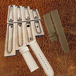 Könnte beinhalten: Ein Satz Metallstanzformen für Uhrenarmbänder auf einem Holzsockel, neben zwei fertigen Uhrenarmbändern. Ein Armband ist olivgrün mit weißen Nähten, das andere ist cremefarben mit blauen und roten Ziernähten.