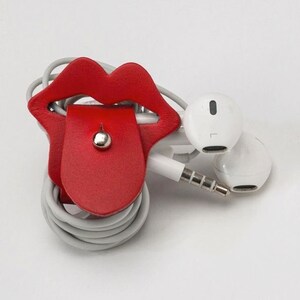 Puede incluir: Porta auriculares de cuero rojo con forma de labios con la lengua fuera. El soporte est&aacute; envuelto alrededor de un par de auriculares blancos.