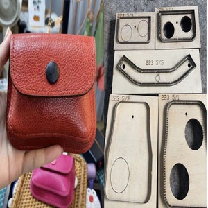 May include: A red leather wallet with a flap closure and a metal button. The wallet is being held in a hand. There are five wooden cutting dies with different shapes and sizes, including a rectangle with two circles, a curved shape, a rectangle with one circle, and a rectangle with two circles. The dies are labelled with numbers and fractions.