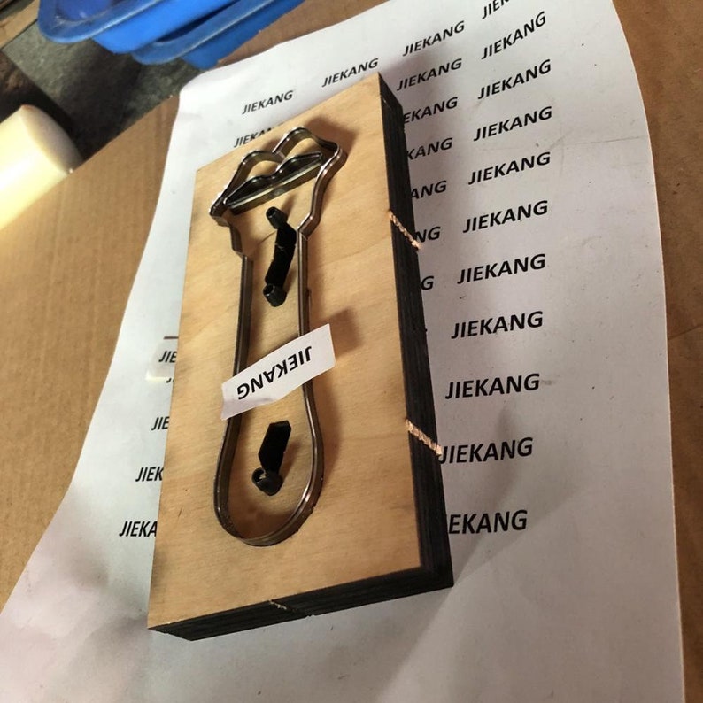 Puede incluir: Una plantilla de troquelado de madera con un recorte de metal en forma de tubo de l&aacute;piz labial. La plantilla est&aacute; etiquetada con "JIEKANG" en el lateral.