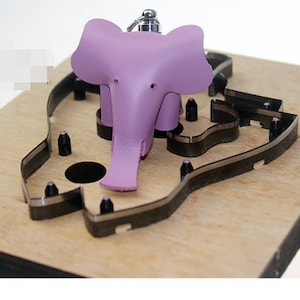 Peut inclure: Un charme en cuir d'éléphant violet avec un anneau métallique attaché au sommet. Le charme est posé sur une planche à découper en bois avec une découpe en métal en forme d'éléphant.