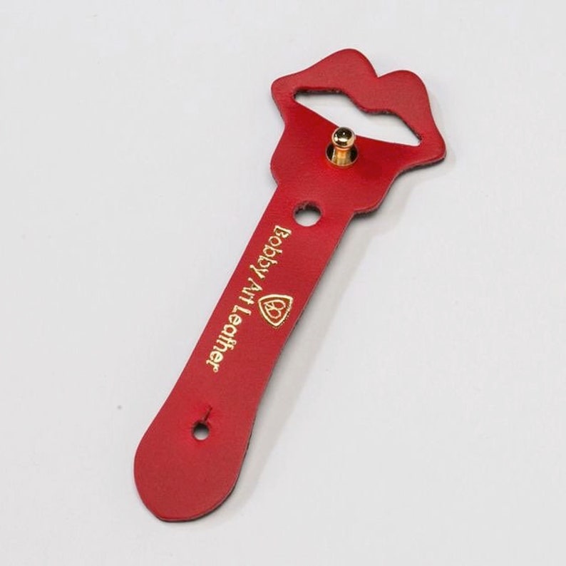 Puede incluir: Llavero de cuero rojo con un logotipo dorado de Bobby Hill Leather y un lazo en forma de labios rojos.