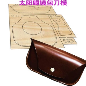 Könnte beinhalten: Ein braunes Lederetui für Brillen mit einem Druckknopfverschluss. Das Etui besteht aus einem einzigen Stück Leder und hat eine geschwungene Form. Ein Schnittmuster für das Etui ist auf einem Stück Holz enthalten.