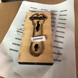 Puede incluir: Una herramienta de troquelado de madera con un recorte de metal de un par de labios y una corbata. La herramienta est&aacute; etiquetada como "JIEKANG".