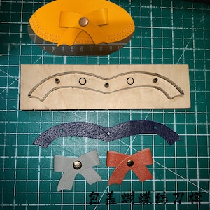 May include: A selection of leather craft components, featuring a yellow bow-shaped pouch, a wooden die, a navy blue leather piece, and two bow-shaped leather pieces in grey and orange. The items are on a green cutting mat.