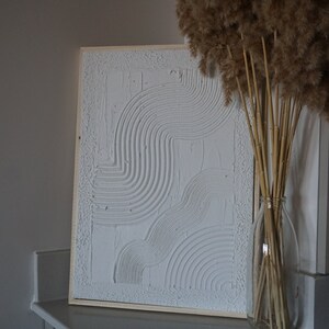 Pode incluir: Obra de arte abstrata texturizada branca emoldurada em madeira. A obra apresenta linhas e formas curvas. Uma garrafa de vidro transparente com capim-dos-pampas seco.