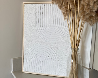 Puede incluir: Un lienzo blanco enmarcado con un diseño abstracto blanco texturizado de dos arcos con líneas. El lienzo está sobre una encimera gris claro con un jarrón de hierba de la pampa seca.