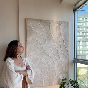 Puede incluir: Gran obra de arte abstracto con una paleta de colores neutros. La obra presenta un diseño texturizado y en capas en tonos beige y blanco, enmarcado en madera natural. La pieza se exhibe en interiores cerca de una ventana.