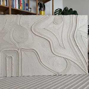 K&ouml;nnte beinhalten: Ein gro&szlig;er wei&szlig;er Wandbehang aus geflochtenem Seil mit einem einzigartigen abstrakten Design mit wirbelnden Mustern und erhabenen Texturen.