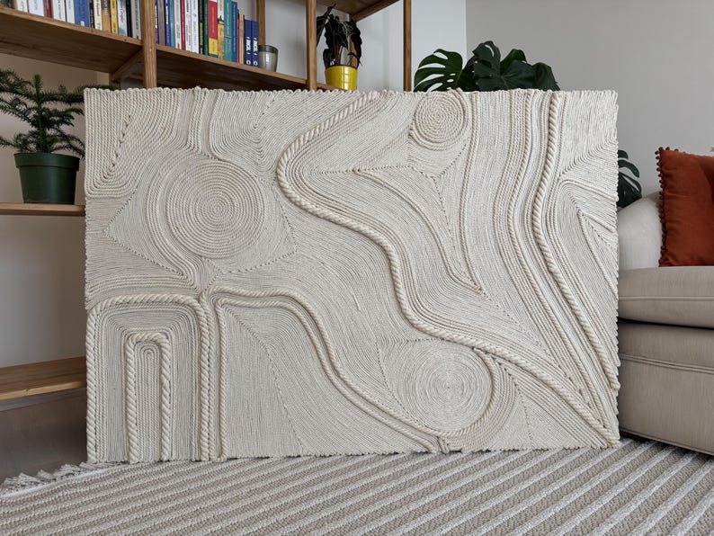 K&ouml;nnte beinhalten: Ein gro&szlig;er wei&szlig;er Wandbehang aus geflochtenem Seil mit einem einzigartigen abstrakten Design mit wirbelnden Mustern und erhabenen Texturen.