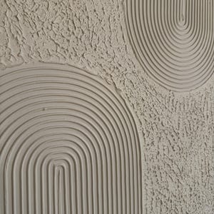 Op de afbeelding: Een gestructureerd wandkunstwerk in een neutrale off-white kleur met verhoogde, uitgesneden boogpatronen en concentrische lijnen. Het oppervlak toont een combinatie van gladde, gegroefde boogvormen en gebieden met een ruwe, onregelmatige pleistertextuur.