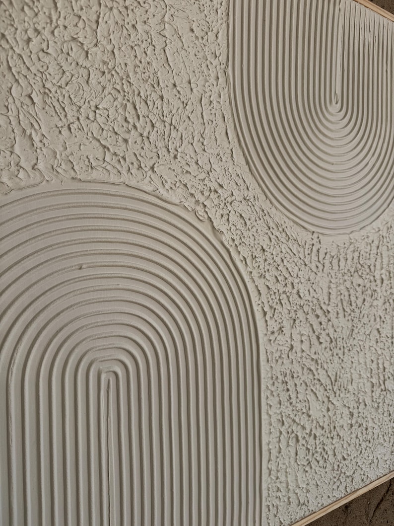 Op de afbeelding: Een gestructureerd wandkunstwerk in een neutrale off-white kleur met verhoogde, uitgesneden boogpatronen en concentrische lijnen. Het oppervlak toont een combinatie van gladde, gegroefde boogvormen en gebieden met een ruwe, onregelmatige pleistertextuur.
