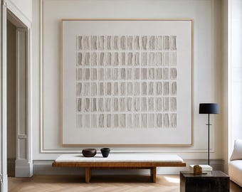 Op de afbeelding: Een groot vierkant wandkunstwerk met een raster van getextureerde lichtgrijze rechthoekige vormen op een witte achtergrond, ingelijst in licht hout. Het kunstwerk hangt boven een lange houten bank met een wit kussen en twee donkere kommen.