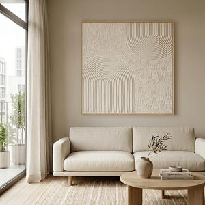 Op de afbeelding: Een vierkant wandkunstwerk met een beige getextureerd oppervlak met verhoogde concentrische boogpatronen, ingelijst in licht hout. Het kunstwerk hangt boven een beige bank, een ronde houten salontafel en een gestreept geweven vloerkleed.