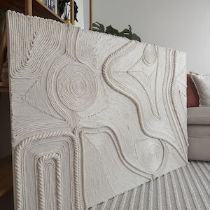 K&ouml;nnte beinhalten: Ein gro&szlig;er, abstrakter Wandbehang aus geflochtenem Seil. Das Seil ist wei&szlig; und erzeugt ein strukturiertes, organisches Muster. Der Wandbehang wird an einer Wand in einem modernen Zuhause ausgestellt.