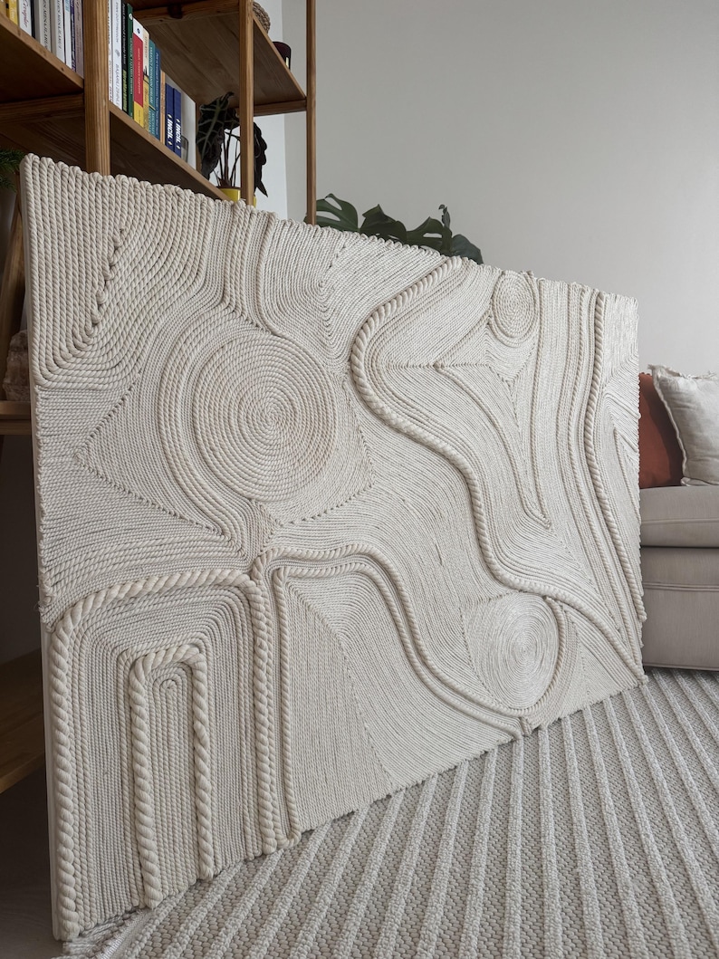 K&ouml;nnte beinhalten: Ein gro&szlig;er, abstrakter Wandbehang aus geflochtenem Seil. Das Seil ist wei&szlig; und erzeugt ein strukturiertes, organisches Muster. Der Wandbehang wird an einer Wand in einem modernen Zuhause ausgestellt.
