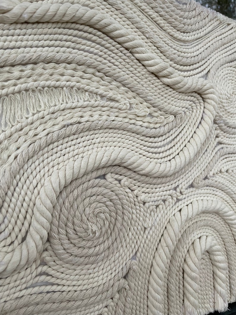 May include: A white rope wall hanging with a swirling pattern. The rope is woven into a series of concentric circles and curves, creating a textured and abstract design.