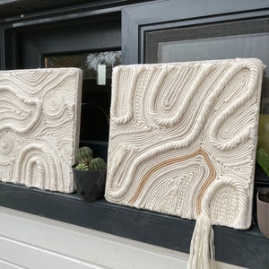May include: Two white macrame wall hangings with a rope design. The hangings are displayed on a windowsill with a small potted plant on each side.