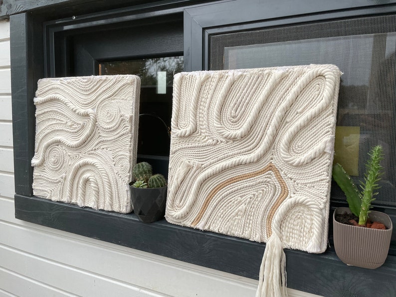 May include: Two white macrame wall hangings with a rope design. The hangings are displayed on a windowsill with a small potted plant on each side.