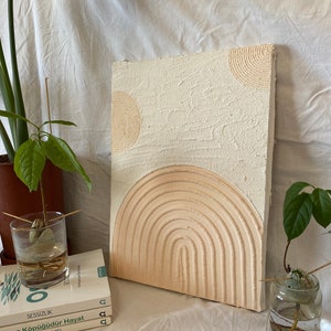 May include: A white textured canvas with a raised, concentric circle design in beige. The design resembles a stylized sun or moon.