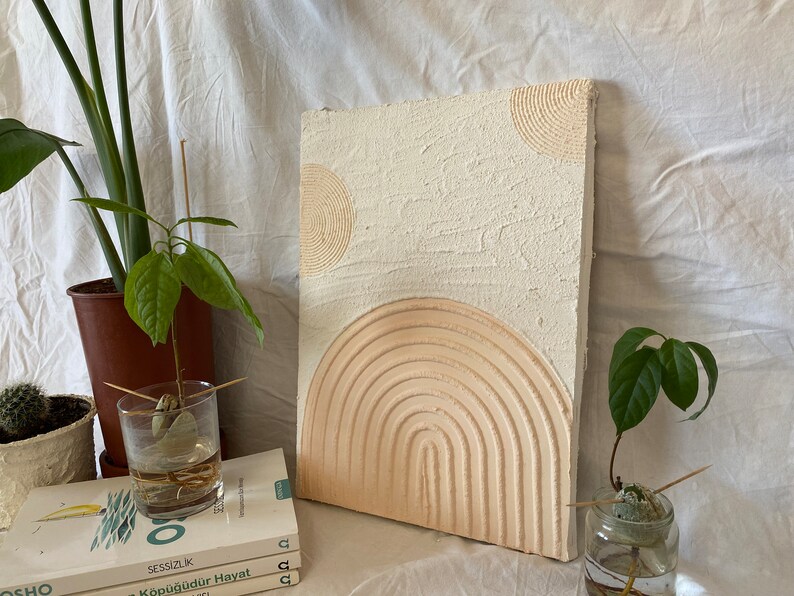 May include: A white textured canvas with a raised, concentric circle design in beige. The design resembles a stylized sun or moon.