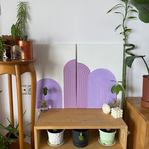 May include: Two purple and white abstract paintings with a textured, raised design. The paintings are hung on a white wall above a wooden shelf with three potted plants.
