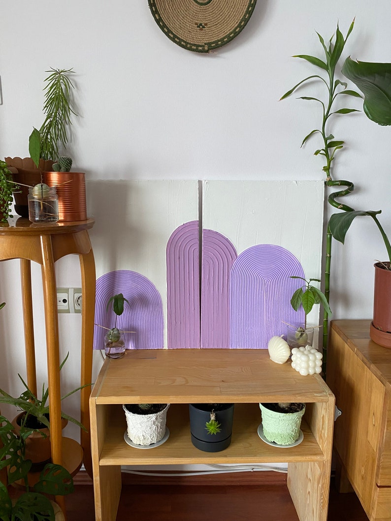 May include: Two purple and white abstract paintings with a textured, raised design. The paintings are hung on a white wall above a wooden shelf with three potted plants.