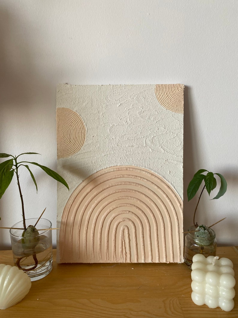 May include: A white textured canvas with a pink raised rainbow design and two beige circles with concentric circles. The canvas is on a wooden surface.