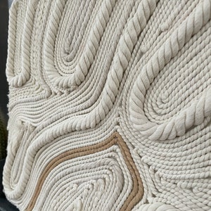 May include: A wall hanging made of woven rope in a cream colour with a brown rope accent. The rope is woven into a pattern of swirls and curves.