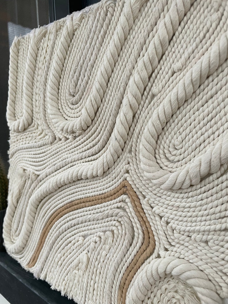 May include: A wall hanging made of woven rope in a cream colour with a brown rope accent. The rope is woven into a pattern of swirls and curves.