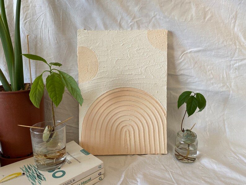 May include: A white canvas with a textured surface and three raised, concentric circles in beige. The circles are arranged in a semi-circle shape. The canvas is leaning against a white surface.
