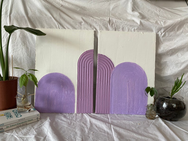 May include: Two white canvases with purple textured arches painted on them. The canvases are leaning against a white surface.
