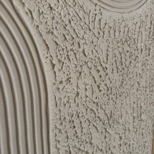 Op de afbeelding: Een gestructureerd wandkunstwerk in een neutraal gebroken wit kleurenpalet. Het oppervlak toont een combinatie van verhoogde, parallelle gebogen lijnen en een dicht, ruw impasto-gedeelte met diepe richels en pieken door dikke verf.