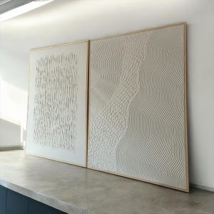 May include: Two white textured abstract paintings with wooden frames. The paintings are hung on a white wall above a gray countertop.