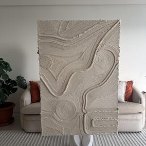 K&ouml;nnte beinhalten: Ein gro&szlig;er beigefarbener Wandbehang aus geflochtenem Seil, der ein strukturiertes, abstraktes Design erzeugt. Der Behang wird von einer Person vor einem beigen Sofa gehalten.