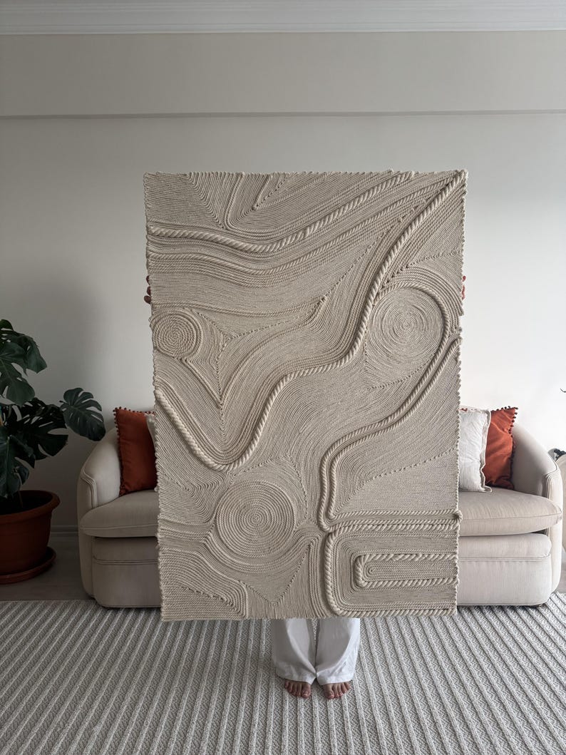 K&ouml;nnte beinhalten: Ein gro&szlig;er beigefarbener Wandbehang aus geflochtenem Seil, der ein strukturiertes, abstraktes Design erzeugt. Der Behang wird von einer Person vor einem beigen Sofa gehalten.