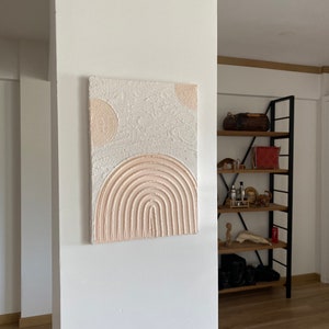 May include: A textured white canvas painting with a pink and white abstract design. The design features a large arch shape with multiple concentric circles and two smaller circles at the top.