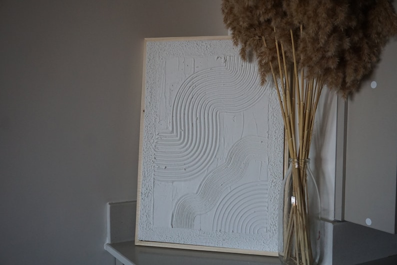 Pode incluir: Obra de arte abstrata texturizada branca emoldurada em madeira. A obra apresenta linhas e formas curvas. Uma garrafa de vidro transparente com capim-dos-pampas seco.